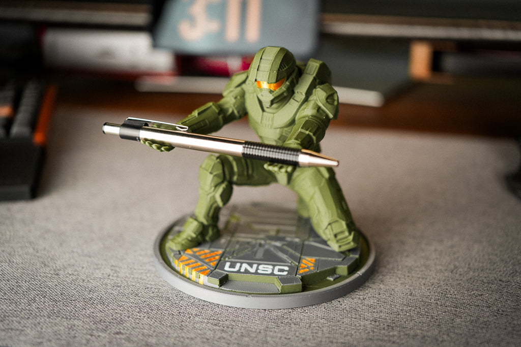 A green UNSC soldier figurine holding a silver and black pen, standing on a round gray base labeled 'UNSC' in a real indoor setting with blurred desk items in the background.