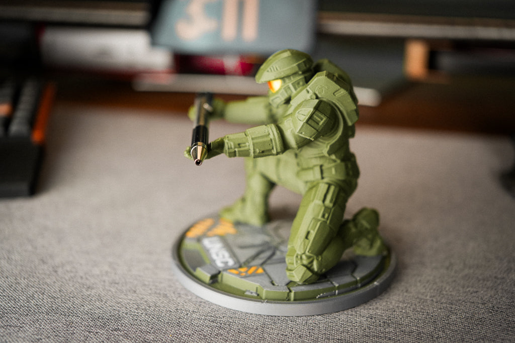 A green sci-fi soldier figurine kneeling on a circular base, holding a rifle, photographed on a gray textured surface with a blurred desk background.