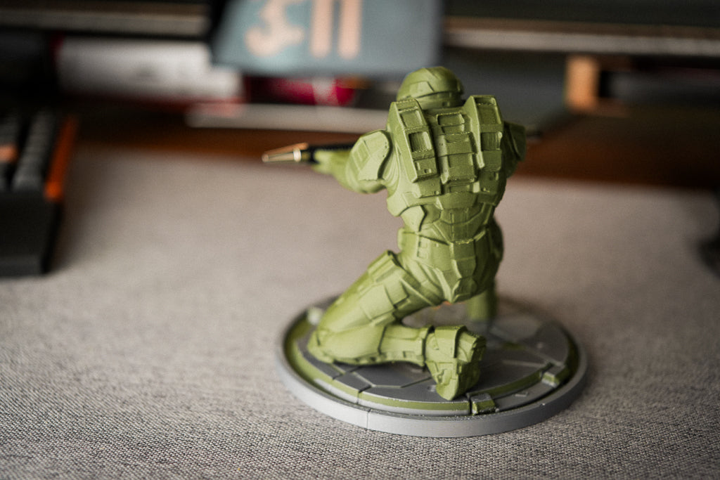 A small olive green miniature figurine of a kneeling soldier on a circular base, photographed on a gray textured surface with a blurred indoor background.