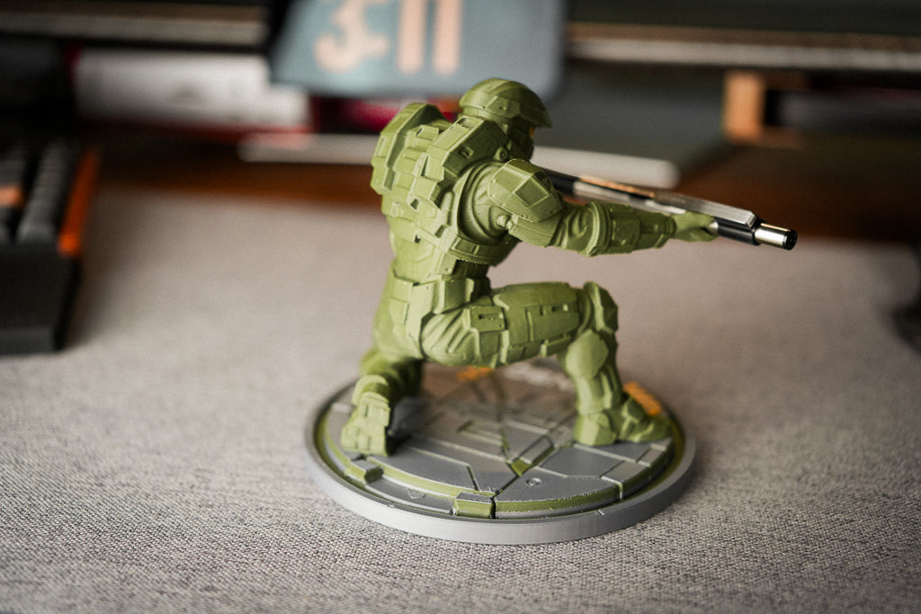 A green sci-fi soldier miniature figurine in a crouching pose, holding a rifle, mounted on a circular gray base. The figurine is photographed on a gray tabletop with a blurred desk background.