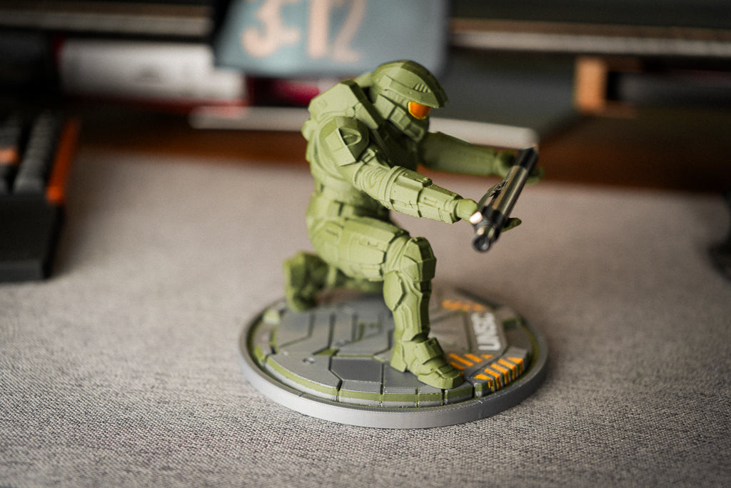 A painted miniature figurine of a kneeling soldier in olive green armor with a black weapon, mounted on a circular gray base, photographed on a gray tabletop with a blurred desk background.
