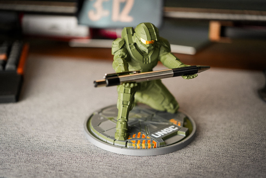 A small green sci-fi armored figurine holding a black and silver pen, mounted on a round gray display base with orange markings, photographed on a gray tabletop with a blurred desk background.