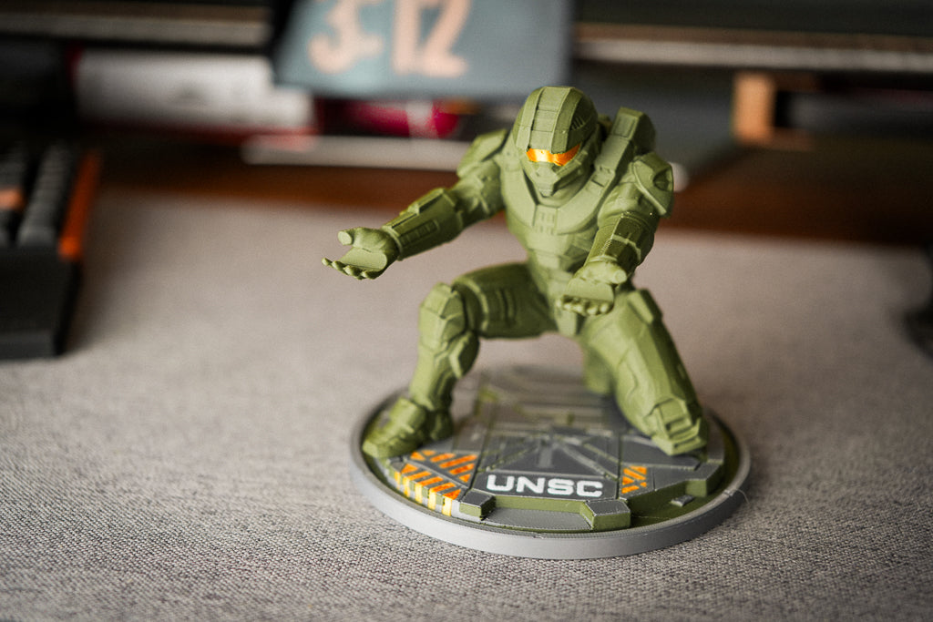 An unpainted green sci-fi mecha figurine posed on a circular gray base with 'UNSC' printed on it, set on a gray tabletop with a blurred desk background.