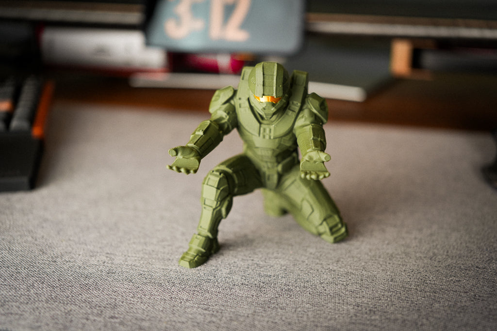 An olive green action figure of a sci-fi warrior in a crouching pose, standing on a gray textured desk with a blurred background showing a keyboard and monitor.