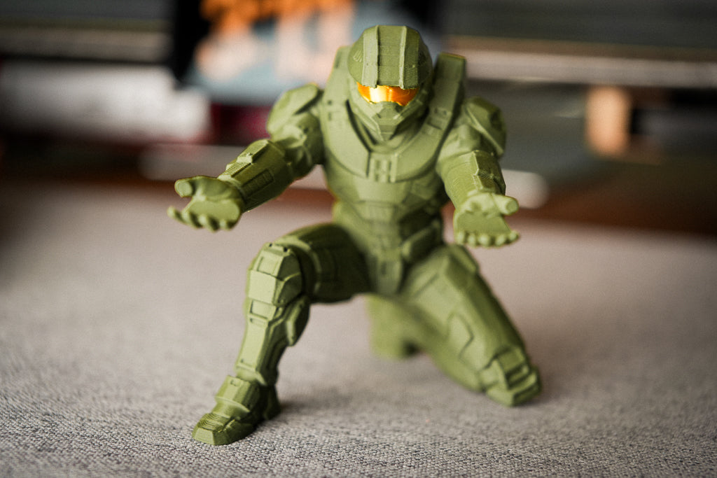 A green Master Chief action figure posed in a crouching stance, featuring a glowing orange visor, set against a blurred indoor background.