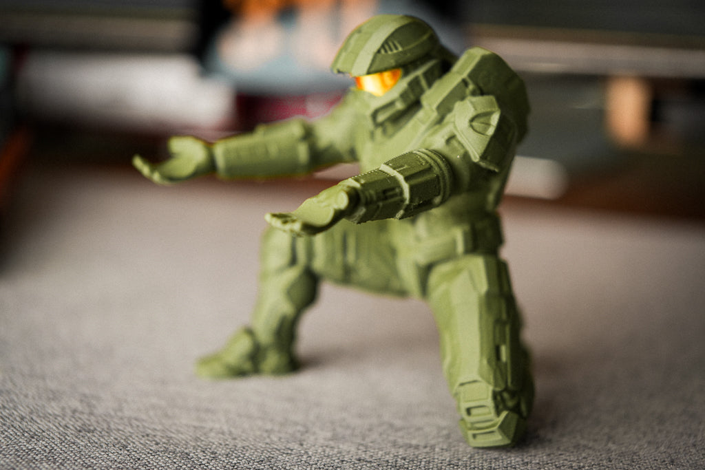 A small green action figure or toy soldier posed in a crouching stance on a gray fabric surface, with a blurred indoor background.