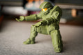 A small green action figure or toy soldier posed in a crouching stance on a gray fabric surface, with a blurred indoor background.