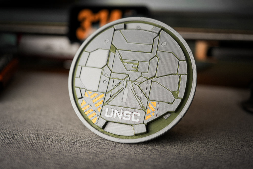 A round UNSC commemorative coin or medallion with a futuristic geometric design, standing upright on a textured gray surface with a blurred background.