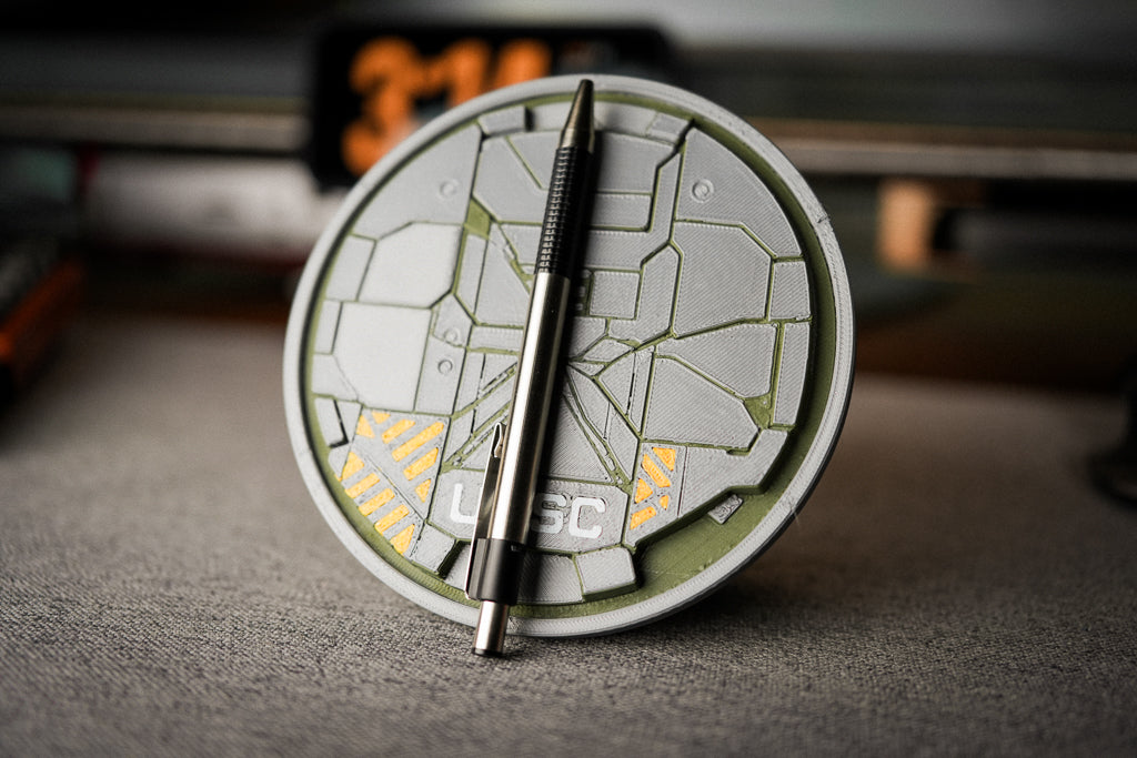 A circular desk clock with a futuristic HUD-style design in gray, olive green, and yellow, featuring a black pen resting on top, set on a gray textured desk surface.