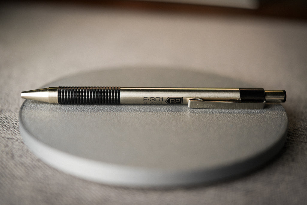 A metallic silver and black retractable pen resting horizontally on a round gray coaster, set against a softly blurred neutral background.